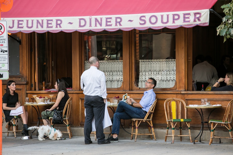 Diners relax with drinks and meals at an outdoor French café with lace-curtained windows and pink awning on a DC street.