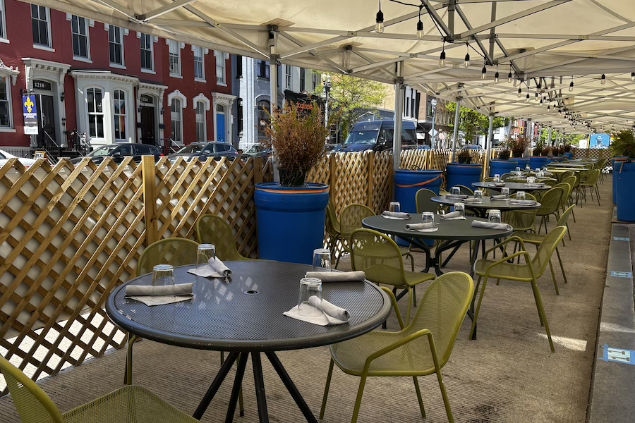 A shaded outdoor dining setup features neatly set green chairs and tables under a canopy along a vibrant urban street.