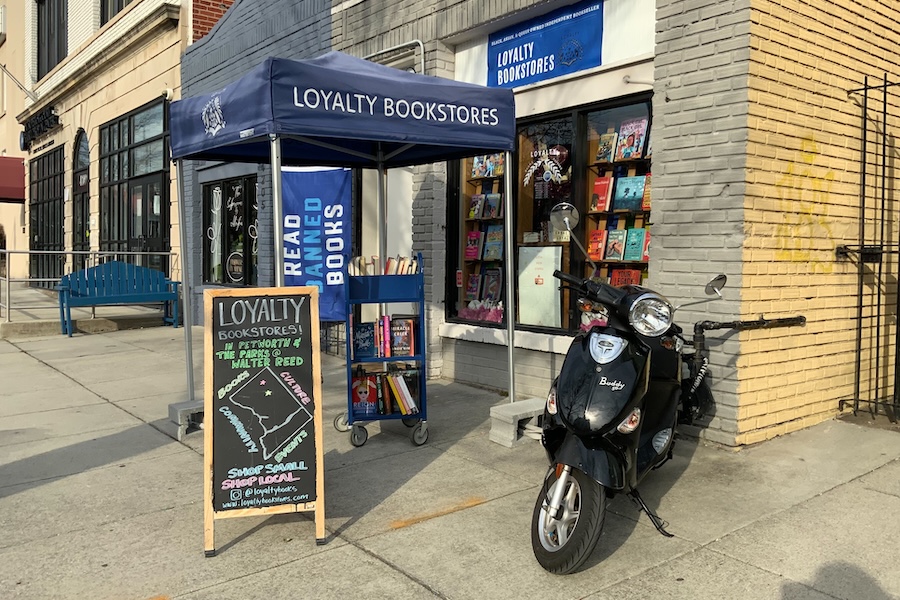 The exterior of Loyalty Bookstores with a blue canopy, book cart, chalkboard sign and scooter parked out front.