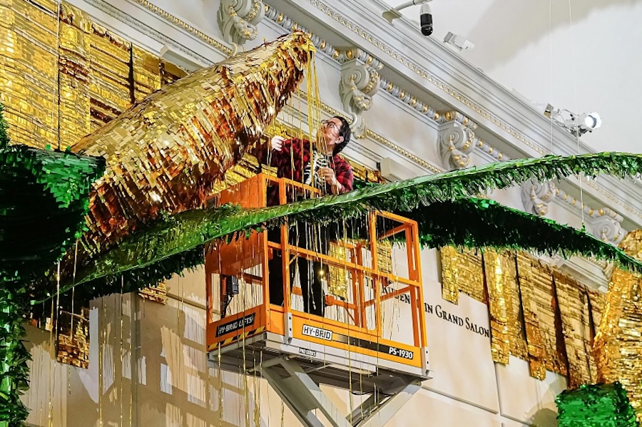 Artist installs a large gold and green piñata-style artwork from a lift inside a grand gallery.