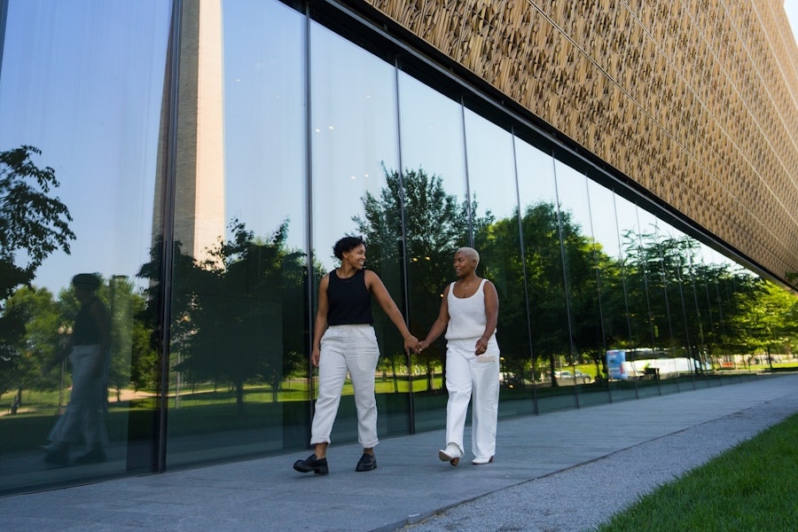 A couple holds hands while walking outside the Smithsonian National Museum of African American History and Culture, with the Washington Monument reflected in the glass.