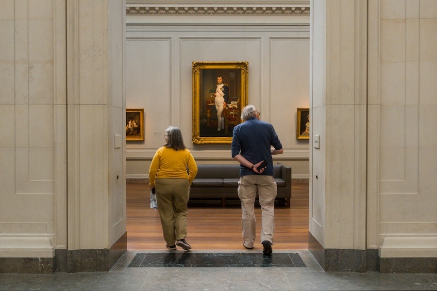Two people walk into a museum gallery toward a large framed portrait hanging on the far wall.