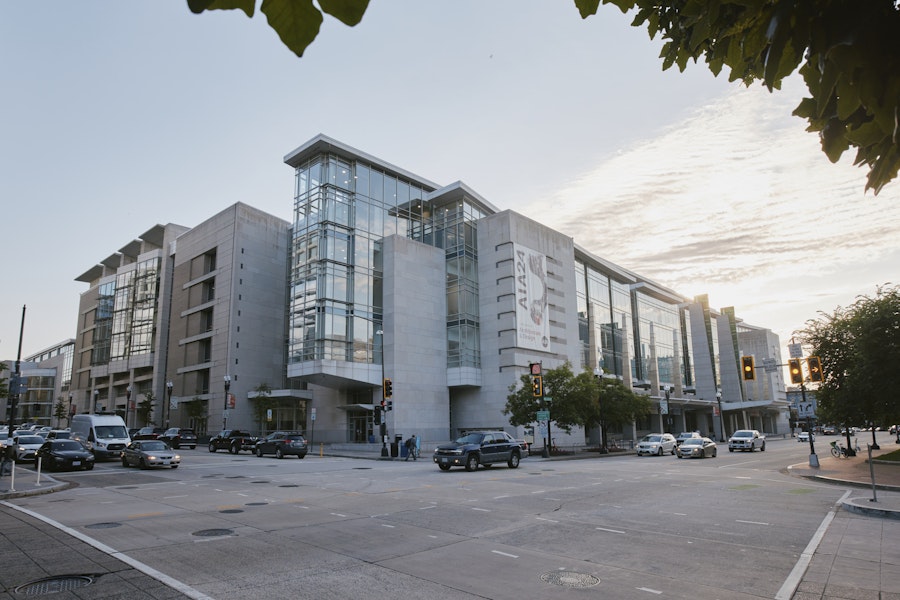 A modern glass and concrete building stands on a city corner with cars parked along the street at dusk.