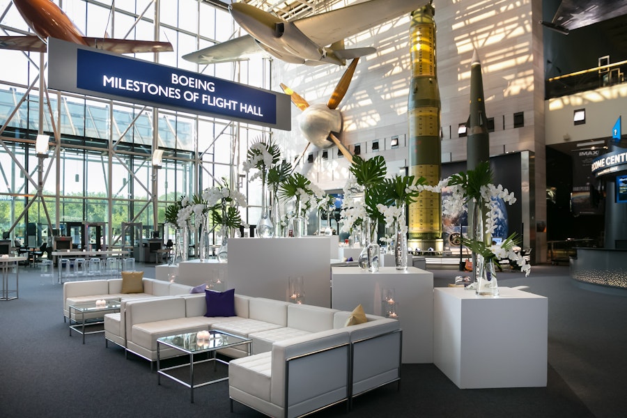 Modern event setup inside the Boeing Milestones of Flight Hall at the Smithsonian National Air and Space Museum, featuring sleek white furniture, floral centerpieces, and historic aircraft suspended overhead.