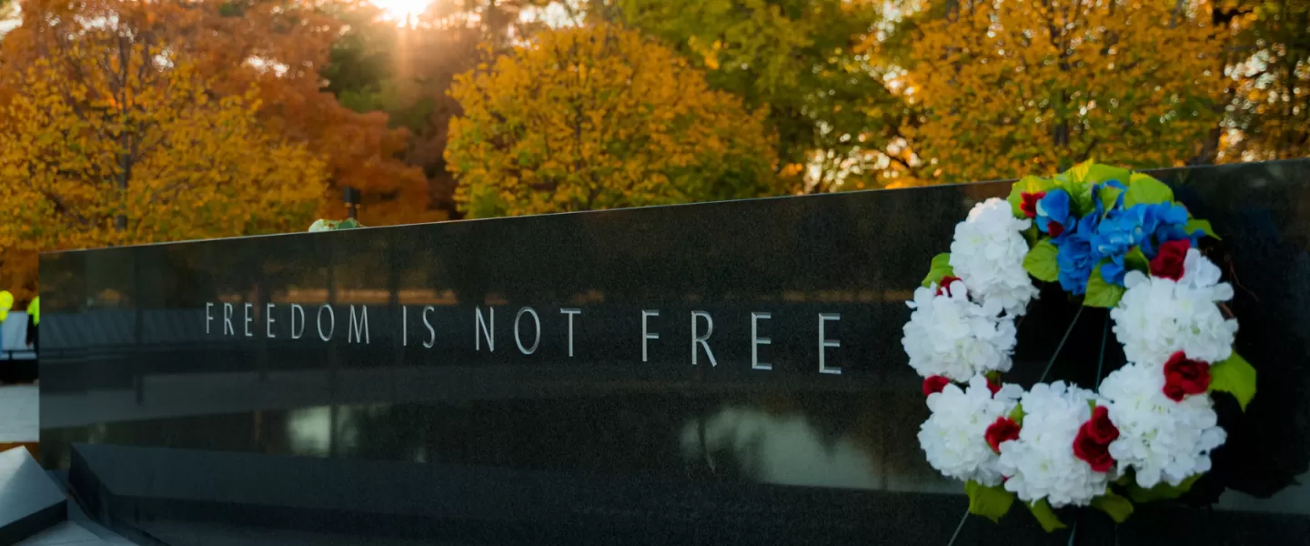 A solemn scene at a memorial, featuring a stone wall engraved with the words "FREEDOM IS NOT FREE." A wreath of red, white, and blue flowers rests against the wall, with autumn trees and the warm glow of a setting sun in the background.