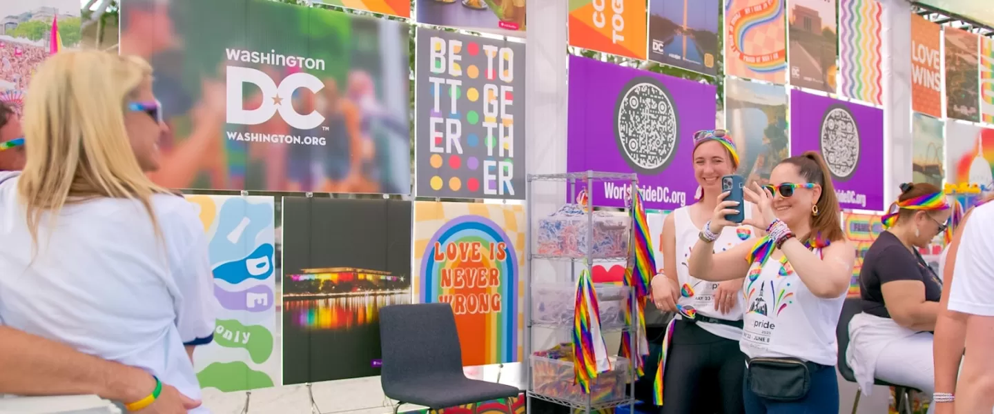 Two people (smiling and dressed in rainbow clothing) take a photo of another person inside a colorful event booth.  person