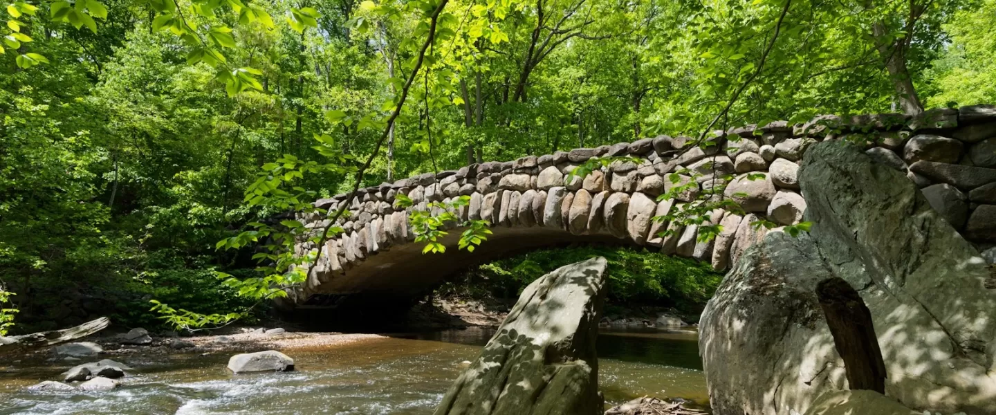A picturesque stone bridge arches over a gently flowing creek surrounded by lush green forest in spring.