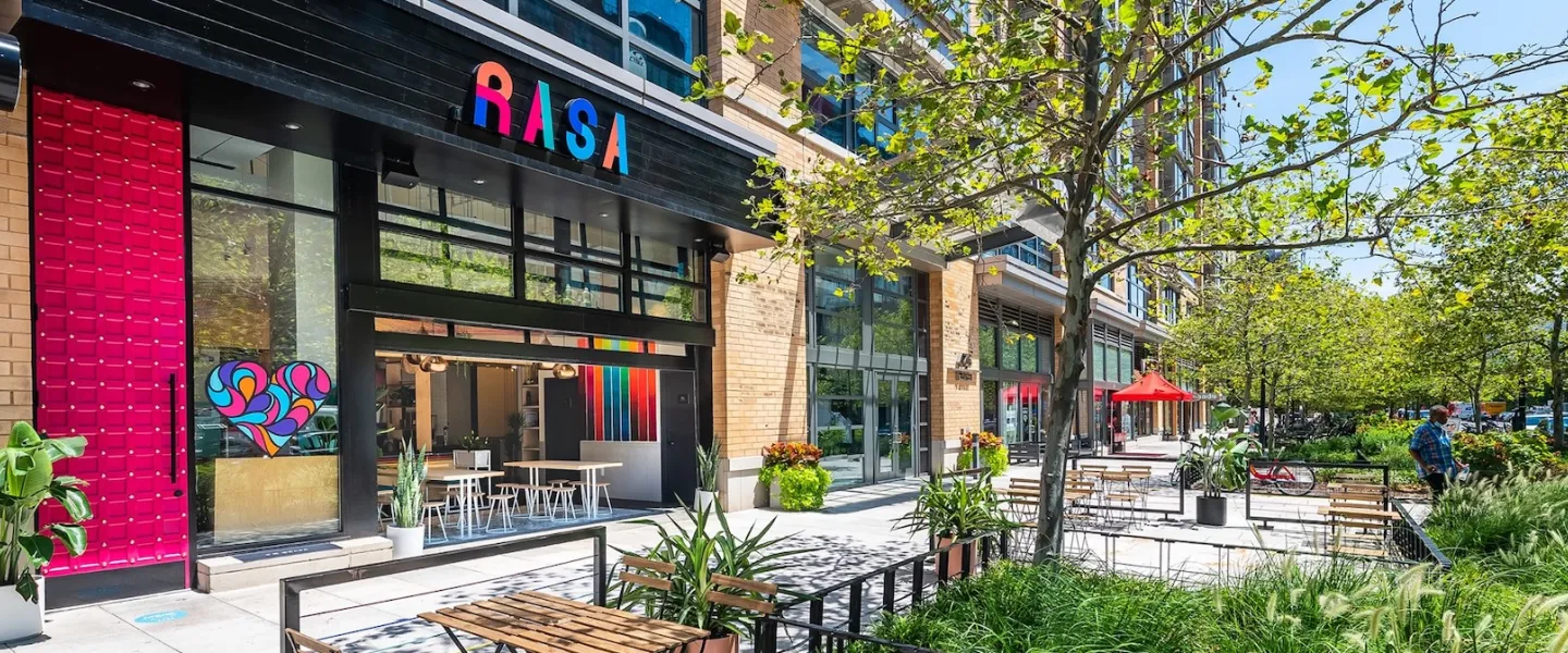 The exterior of Rasa restaurant in Mount Vernon Square features bright modern decor and a sunny outdoor patio with seating and greenery.