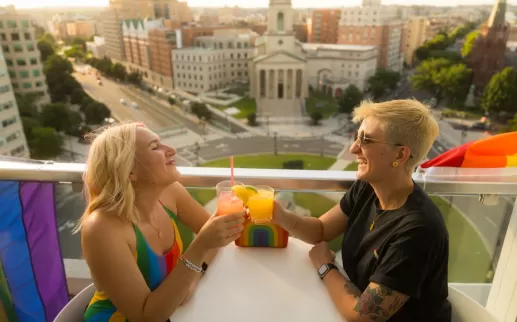 A couple enjoying a cocktail on a rooftop with a view of historic city buildings below. 
