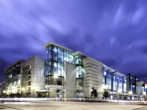 The image shows the exterior of the Walter E. Washington Convention Center in Washington, DC, illuminated at night. The sleek, modern architecture is highlighted by large glass windows and bright lighting, set against a dramatic sky. This convention center is a key venue for conferences, events, and exhibitions in the heart of the nation's capital.