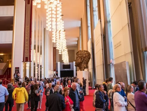 A crowded lobby inside the Kennedy Center, with people walking past a large sculpture and chandeliers under a high ceiling.