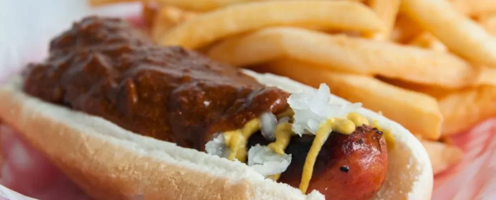 This image shows a chili half-smoke from Ben's Chili Bowl, a DC classic featuring a grilled sausage topped with chili, mustard, and onions, served with fries.