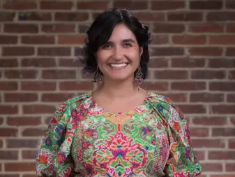 A person with short black hair, wearing a colorful patterned dress, smiling in front of a brick wall.